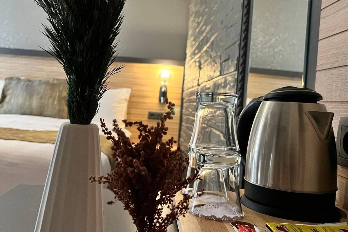 A modern hotel room with a wooden table, a white vase with dried flowers, a clear glass, a stainless steel kettle, and a remote control.