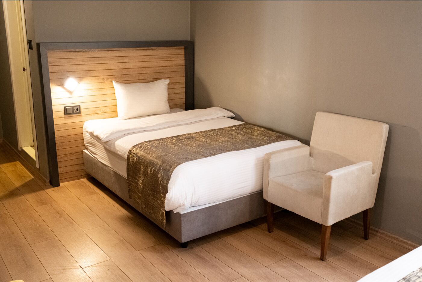 A single bed with a headboard, a bedside table, and a chair in a room with wooden flooring and a neutral wall color.