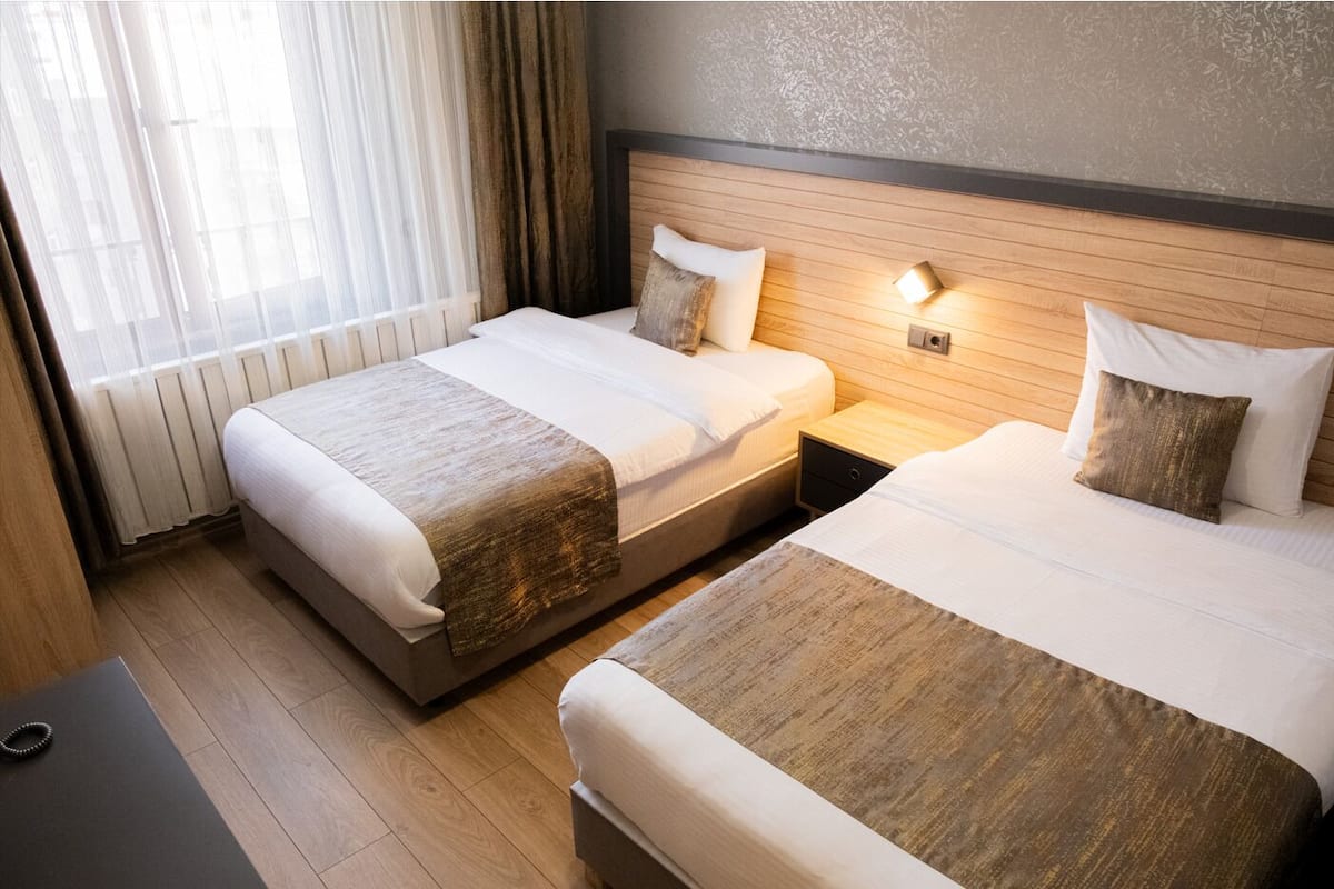 A hotel room with two beds, a wooden headboard, a bedside table with a lamp, and a window with curtains.