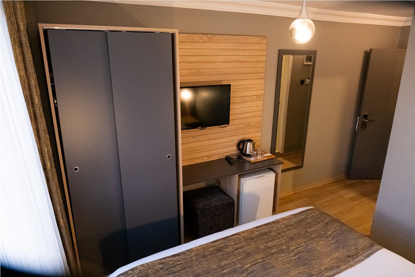 Una habitación de hotel con cama, televisión instalada en un panel de madera, un refrigerador pequeño y un espejo.