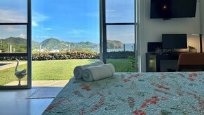 Une chambre d’hôtel dotée d’une grande porte-fenêtre coulissante donnant sur un balcon avec vue sur la mer et les collines. On y trouve un lit recouvert d’une courtepointe à motifs floraux, un bureau avec un ordinateur et un téléviseur fixé au mur.