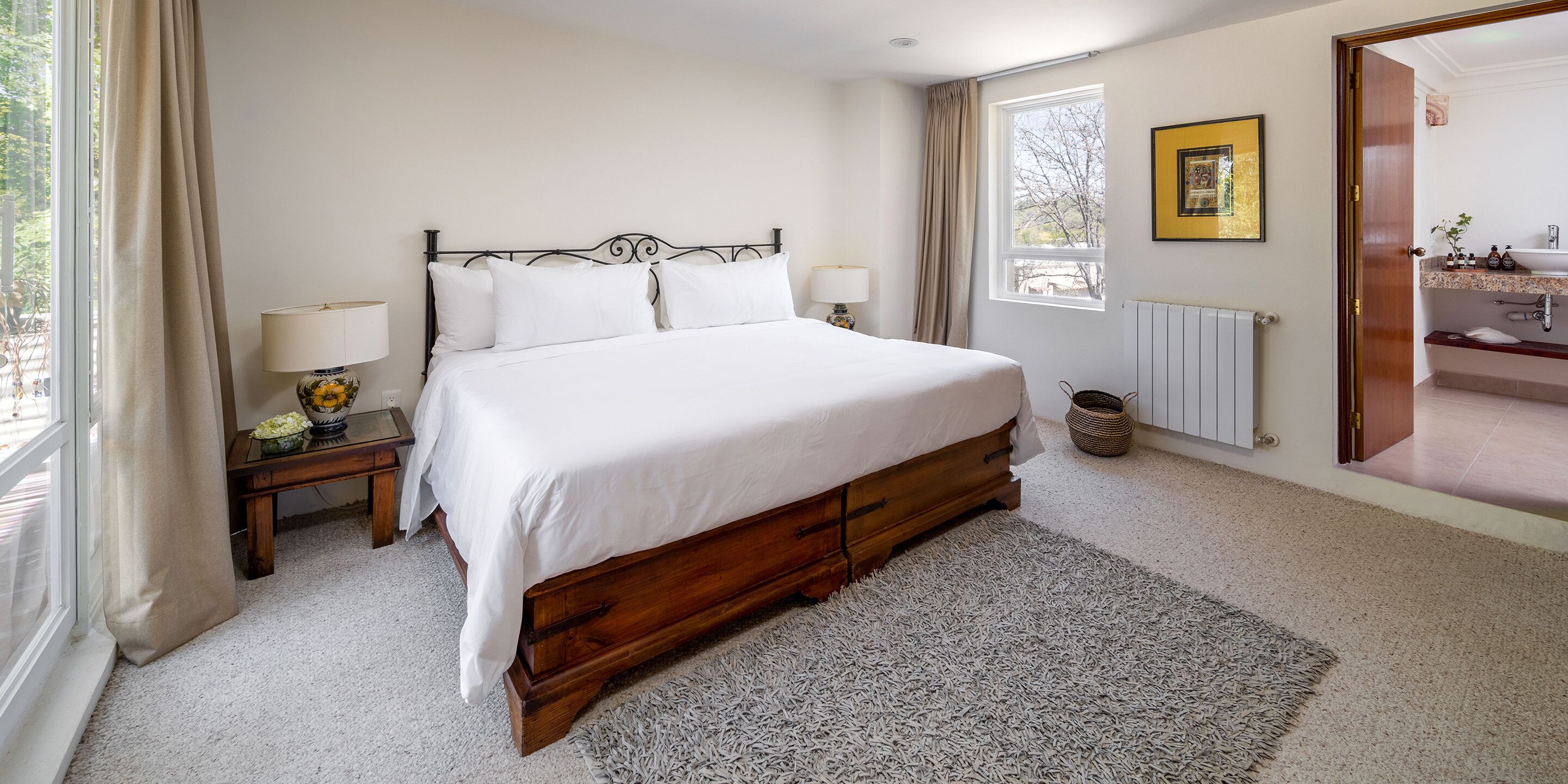 A bedroom with a large bed, a bedside table with a lamp, a window with curtains, and a bathroom visible through an open door.