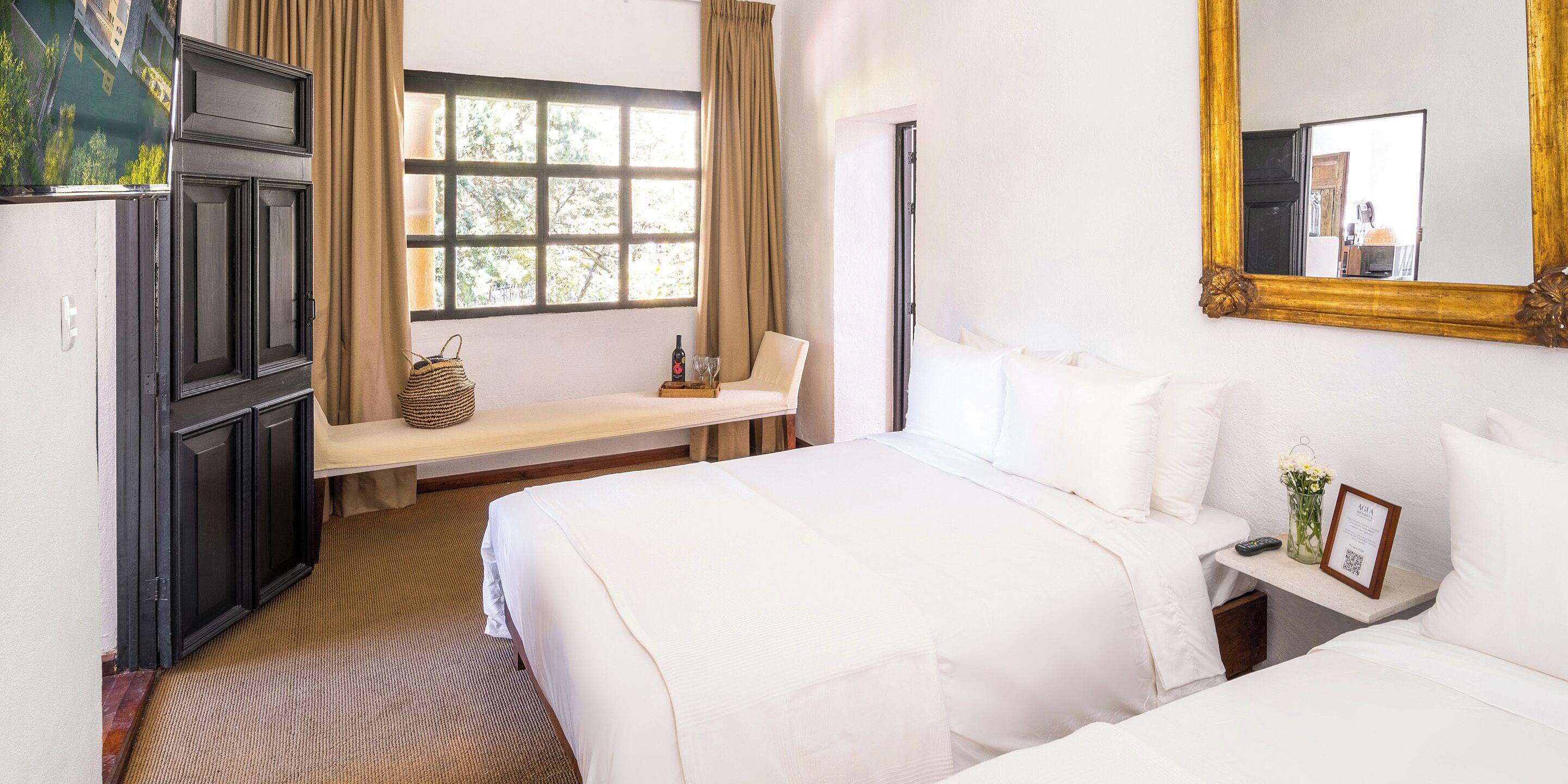 Ein Hotelzimmer mit einem großen Bett, einer Bank mit einem gewebten Korb, einem kleinen Tisch mit einer Vase und einem Spiegel an der Wand.