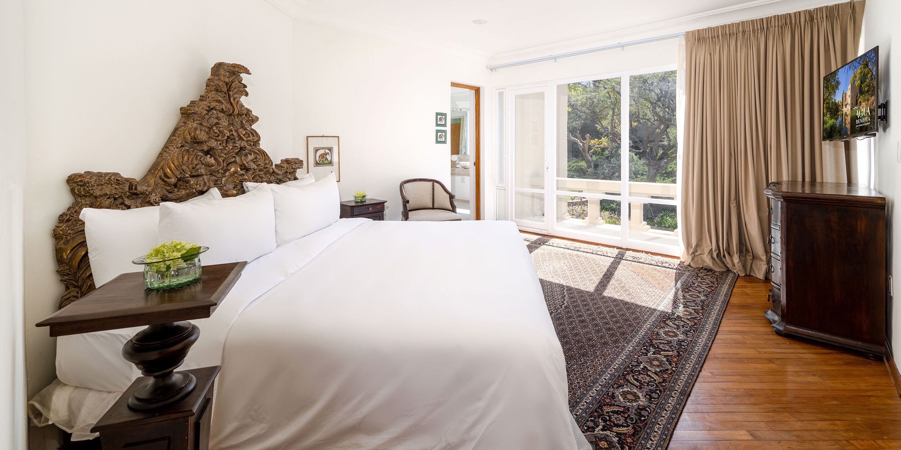 Un dormitorio con una cama grande, un cabecero de madera, un televisor montado en la pared y vistas a zonas verdes por la ventana.