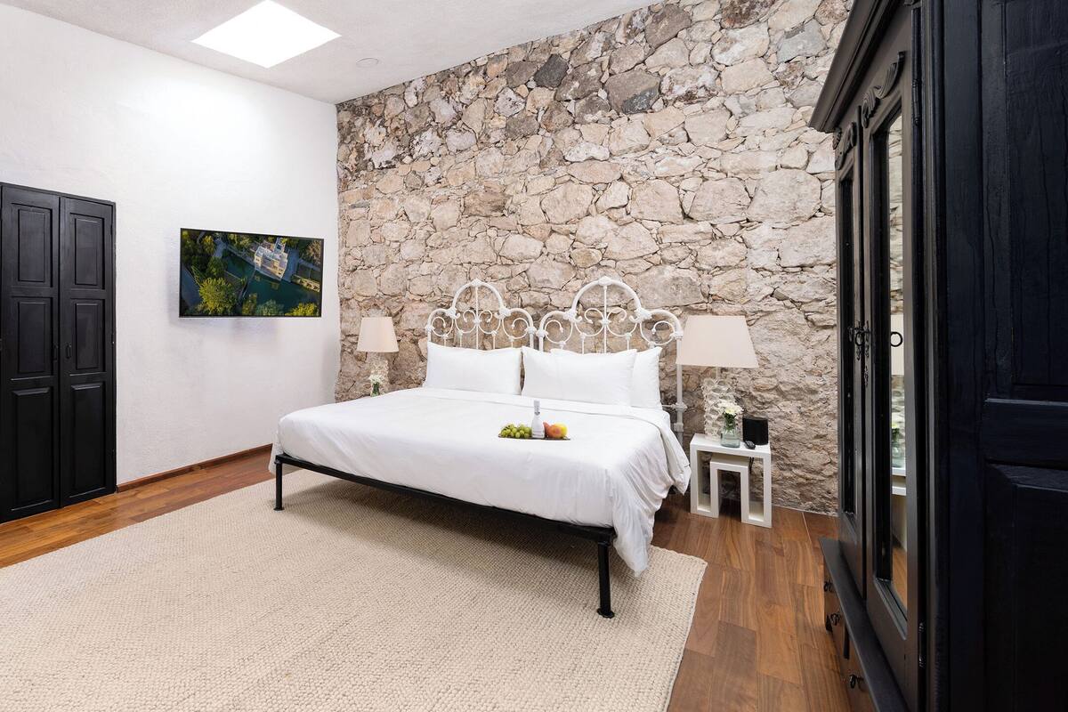 A bedroom with a stone wall, a bed with white linens, a black wardrobe, and a painting on the wall.
