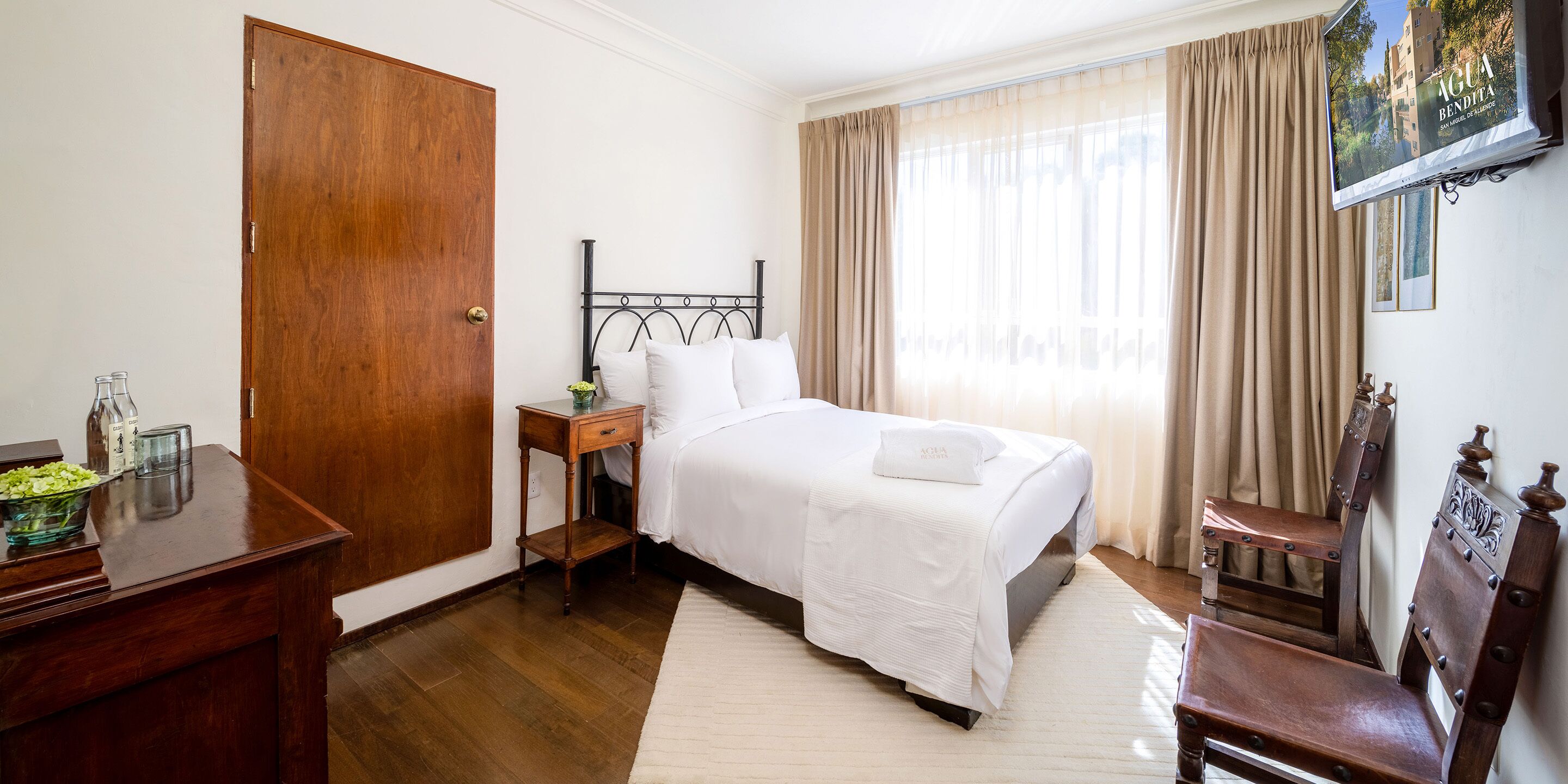 Quarto com porta de madeira, cama com roupa de cama branca, mesa de cabeceira, uma cadeira e janela com cortinas.