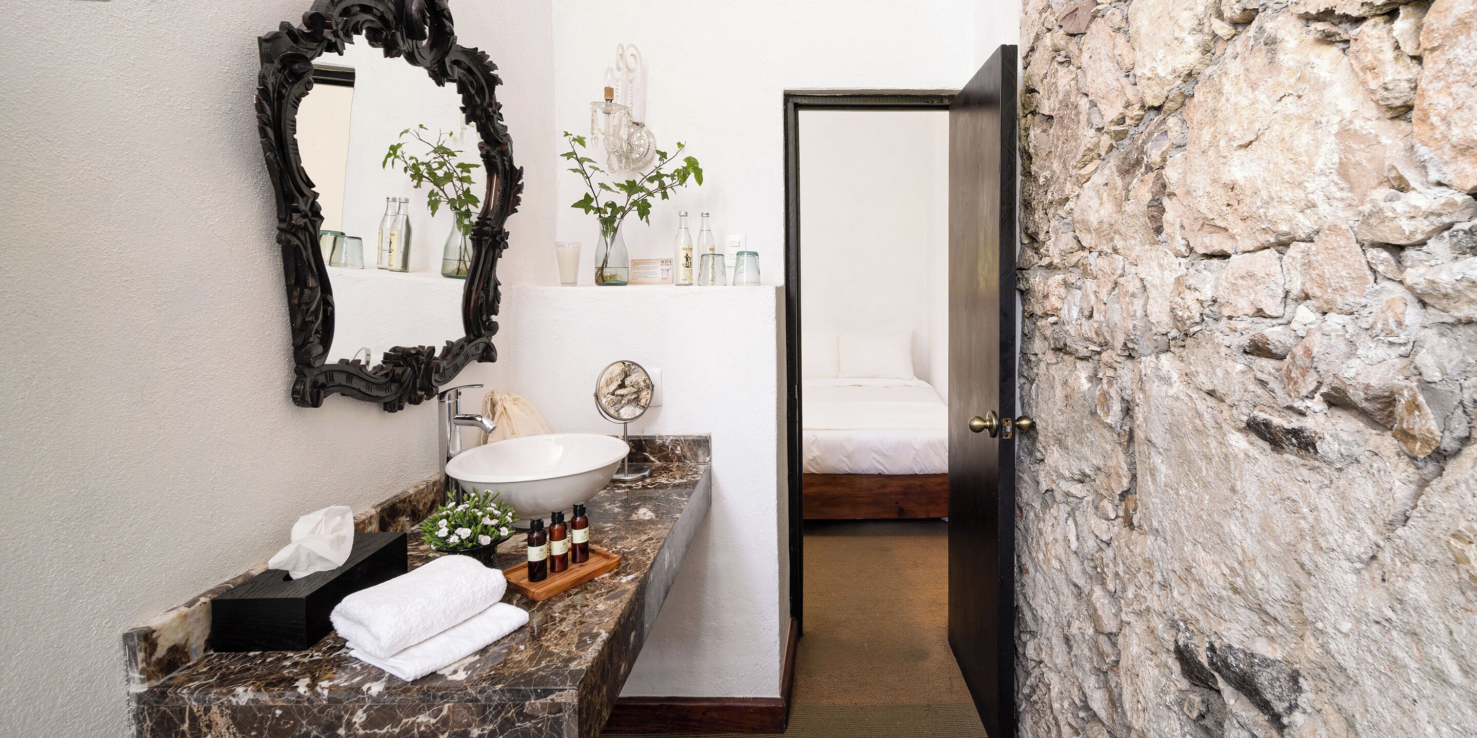 A bathroom with a marble countertop, a white sink, a large mirror, and a bed in the background.
