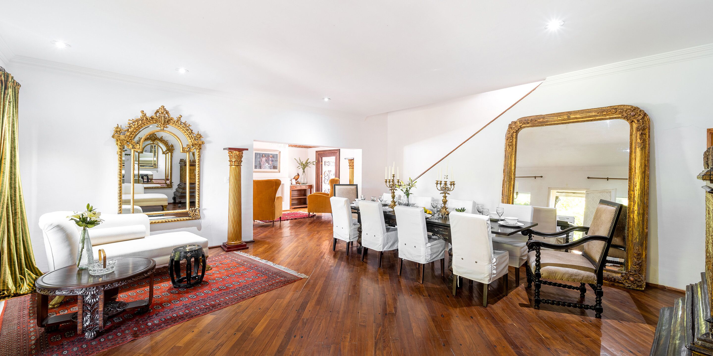 A spacious living room with a dining area, featuring a large ornate mirror, white furniture, and a red patterned rug.