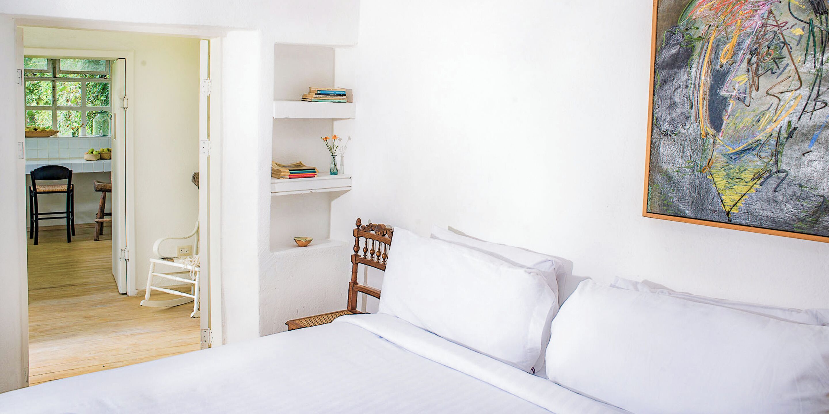 A bedroom with a bed, a wooden chair, a desk with a chair, and a shelf with books and a vase.