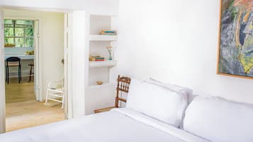 A bedroom with a bed, a wooden chair, a desk with a chair, and a shelf with books and a vase.
