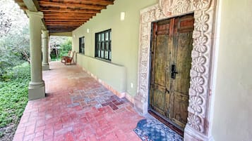 A covered walkway with a wooden ceiling, a large ornate door, and a brick floor leading to a building with a light-colored exterior and decorative floral carvings.