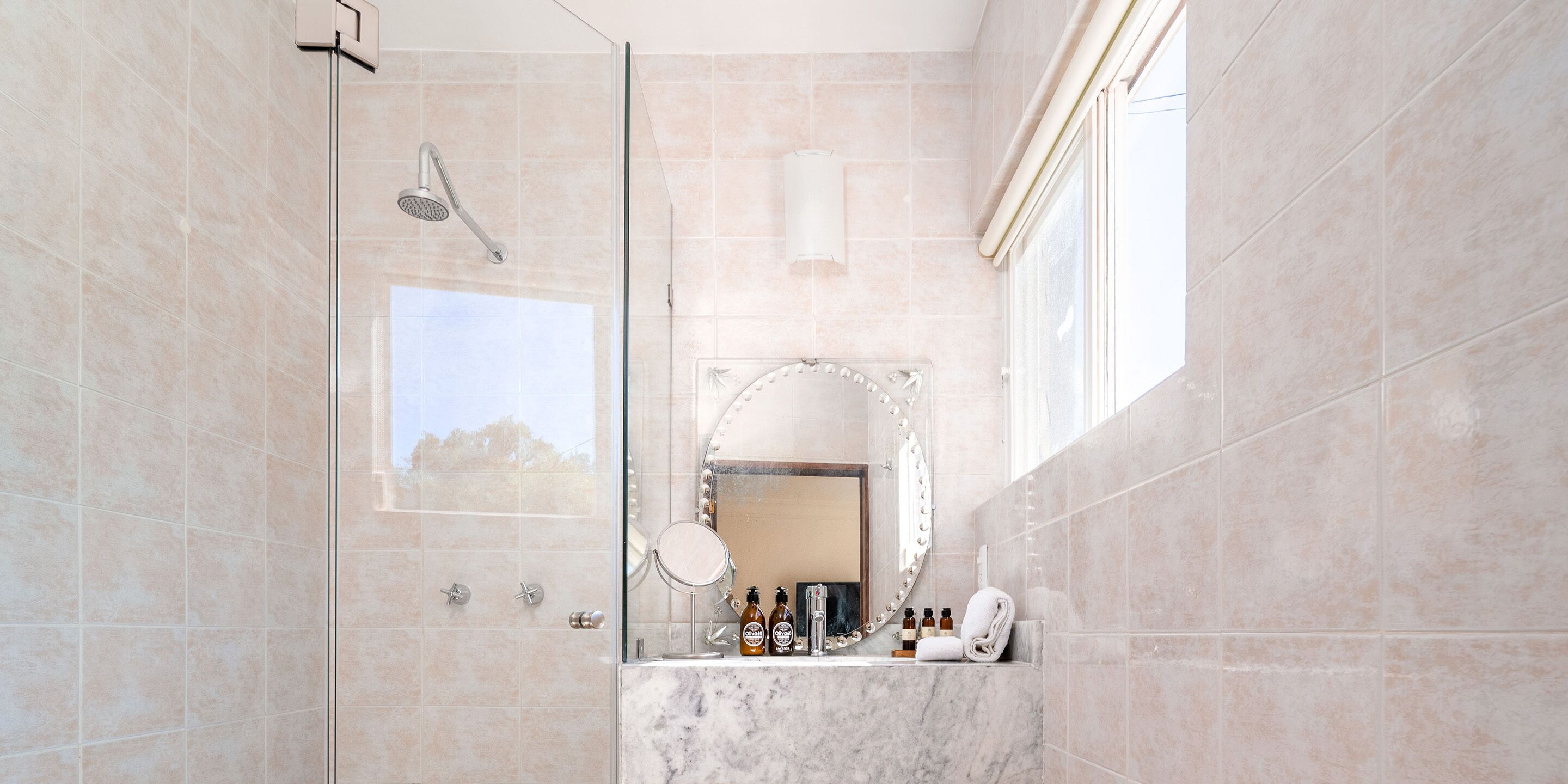A bathroom with a glass-enclosed shower, a marble sink, a vanity mirror, and toiletries.