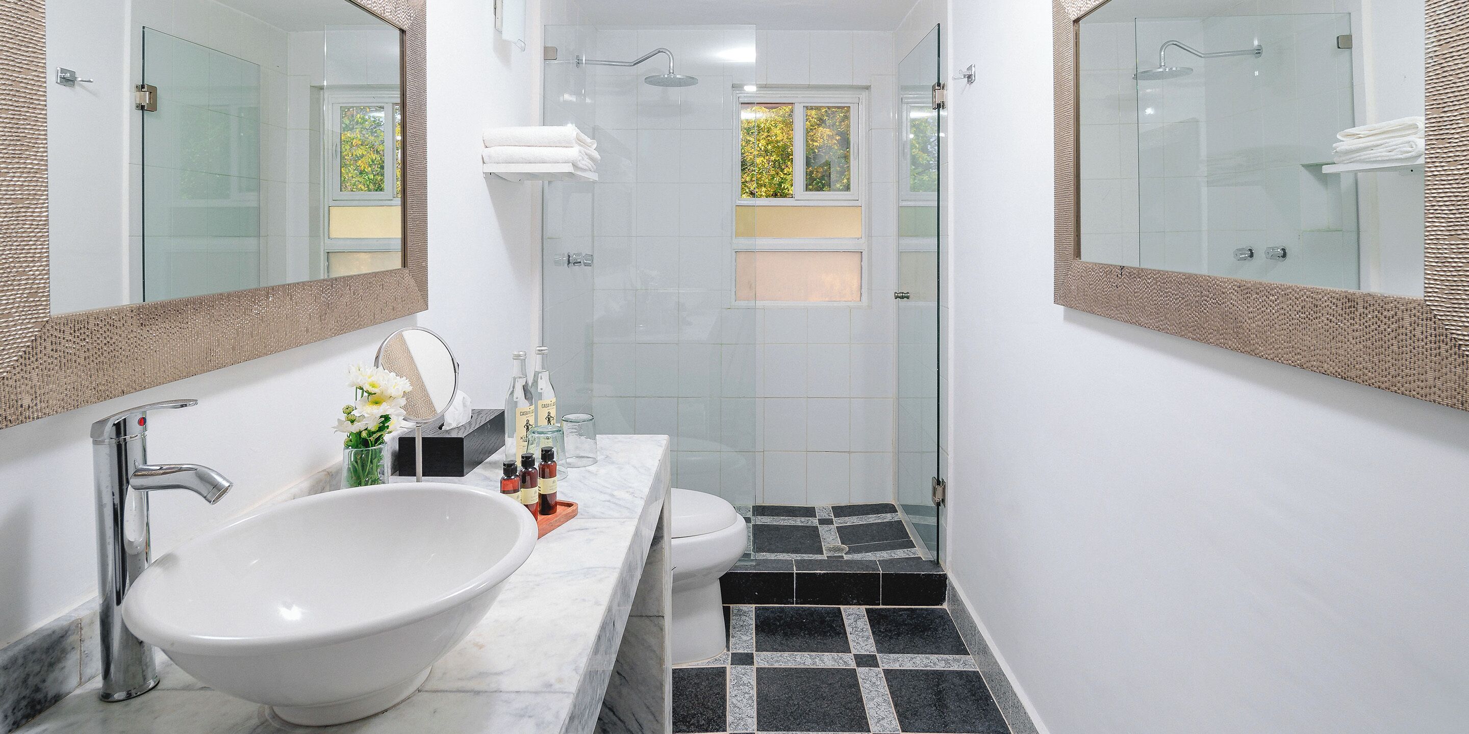 A modern bathroom with a large white sink, two mirrors, a glass shower enclosure, and a black and white tiled floor.