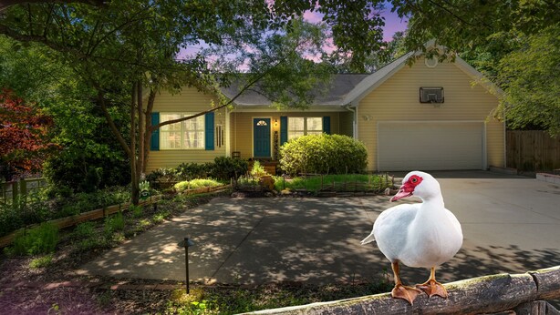Una casa amarilla con jardín delantero, entrada para coches y un pato sobre una valla de madera.