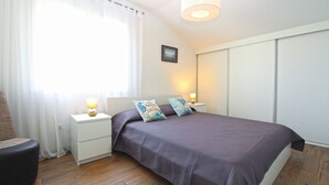 A bedroom with a bed, two bedside tables with lamps, a chair, a window with curtains, and a picture on the wall.