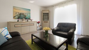 A modern living room with a glass coffee table, two black leather sofas, a beige cabinet, and a painting of a pier.