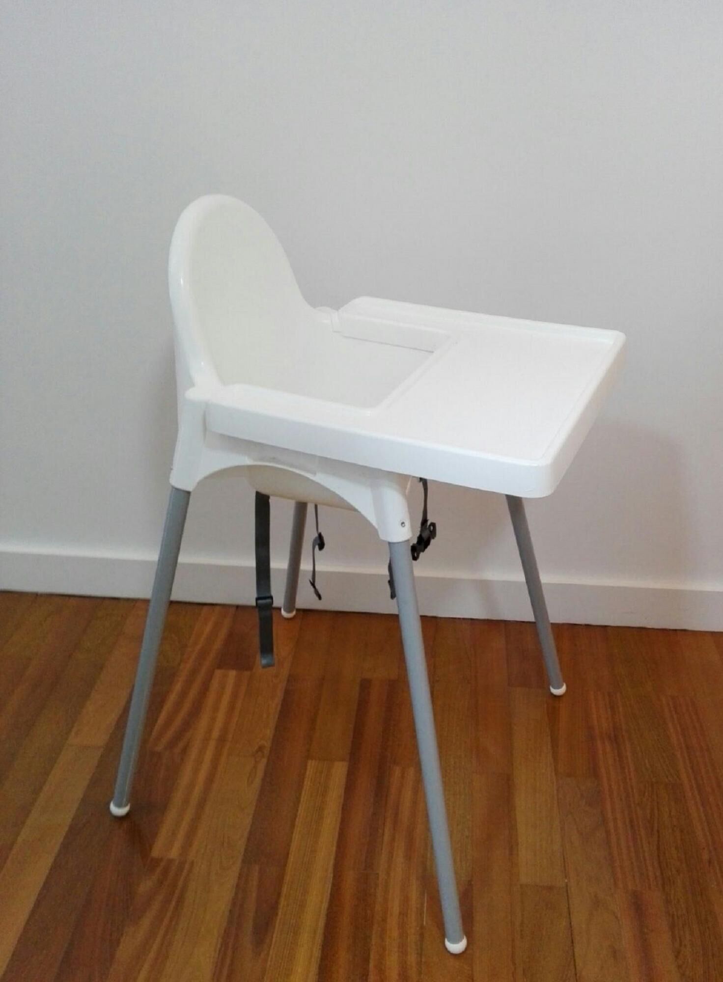 A white high chair with grey legs and a tray table, positioned against a white wall on a wooden floor.