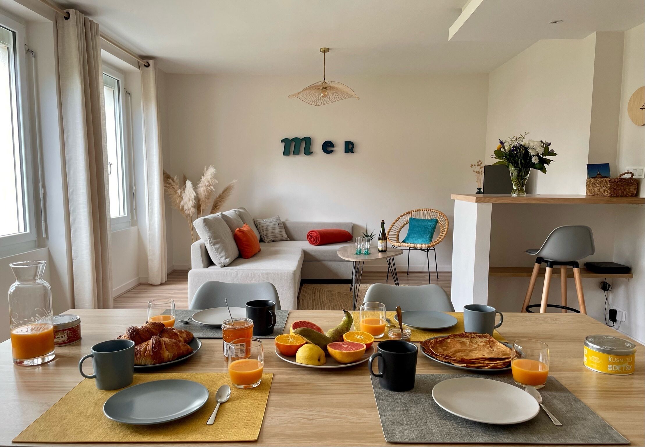 A modern living room with a sofa, a dining table set for breakfast, and a wall with the word 'mer' partially visible.