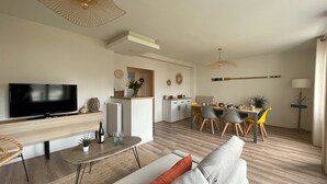 A modern living room with a flat-screen TV, a dining area with a long table and chairs, and a kitchen area with a built-in oven and microwave. - BLEU COMME MER - T2 apartment just a stone's throw from the beach (Concarneau)