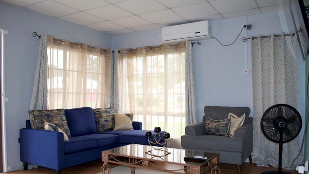 A living room with a blue sofa, a grey armchair, and a coffee table. There is a fan and an air conditioning unit on the wall.