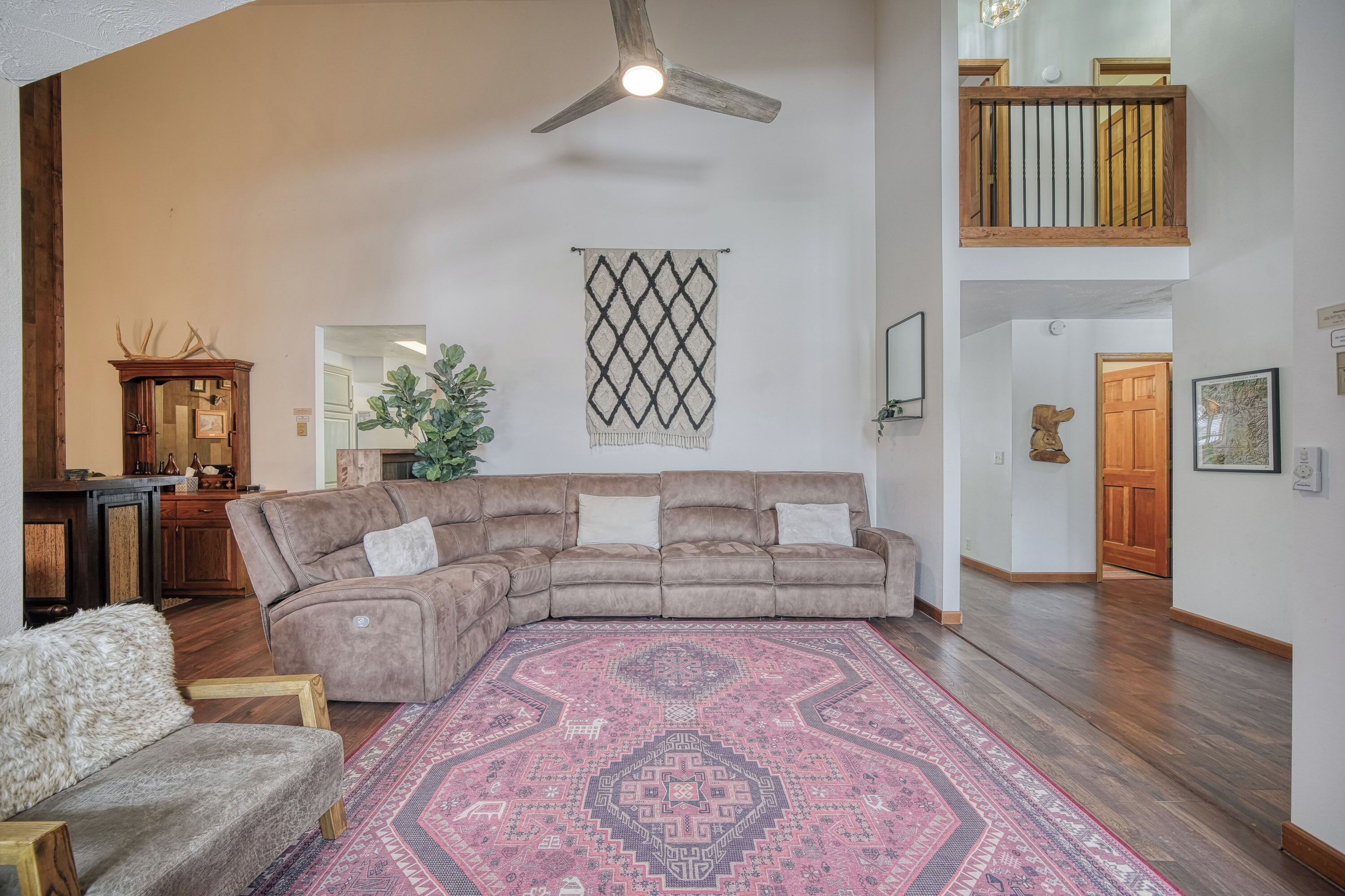 A spacious living room with a large sectional sofa, a wooden ceiling fan, a decorative wall hanging, and a staircase leading to an upper level.