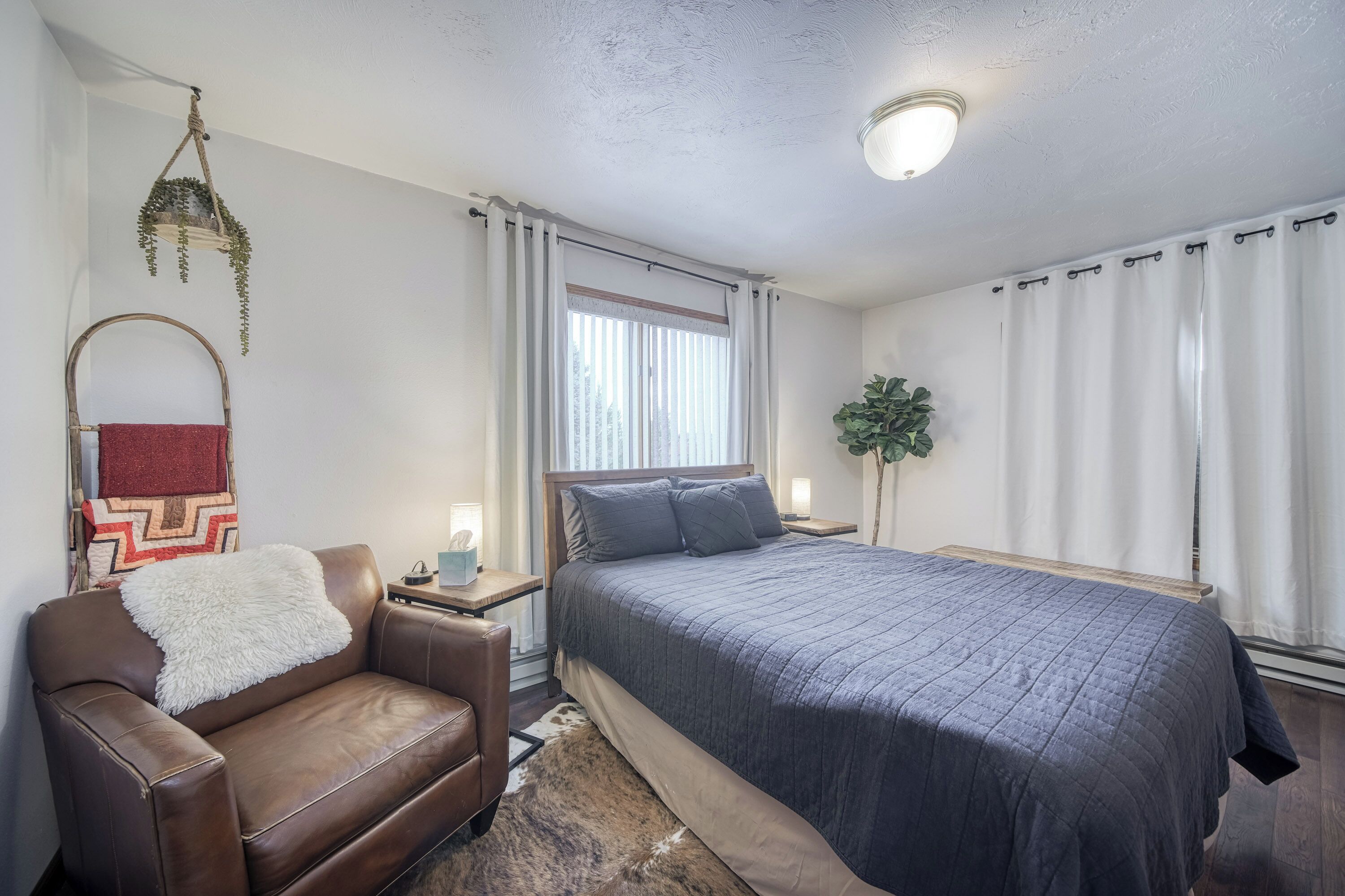 A bedroom with a bed, a leather chair, a hanging plant, and a nightstand with a lamp.