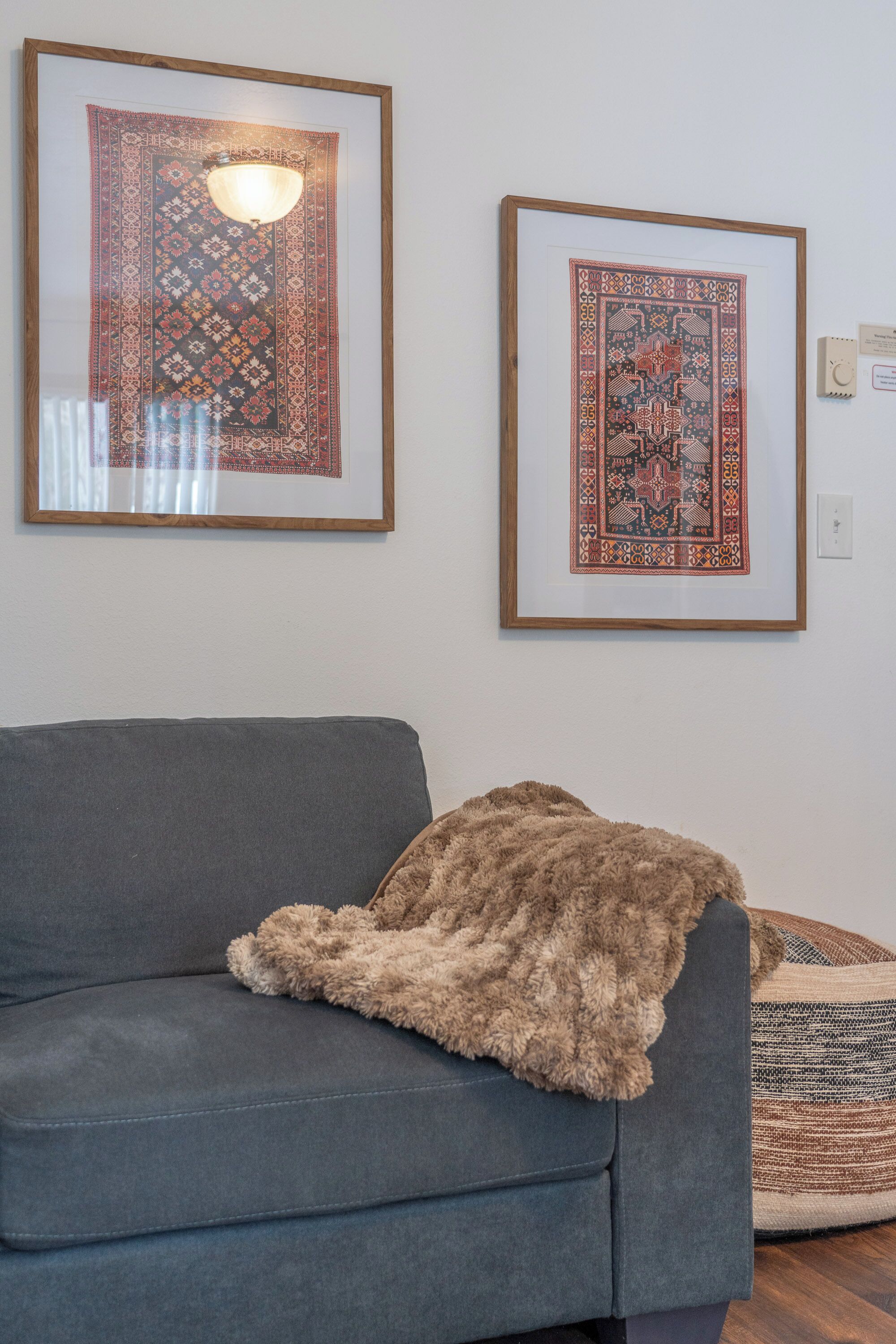 A room with a grey sofa, a brown throw blanket, and two framed rugs on the wall.