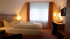 A hotel room with a bed, two bedside lamps, a desk with a chair, a television, and a large window with curtains. - Hotel Jägerhof (Bad Hersfeld)