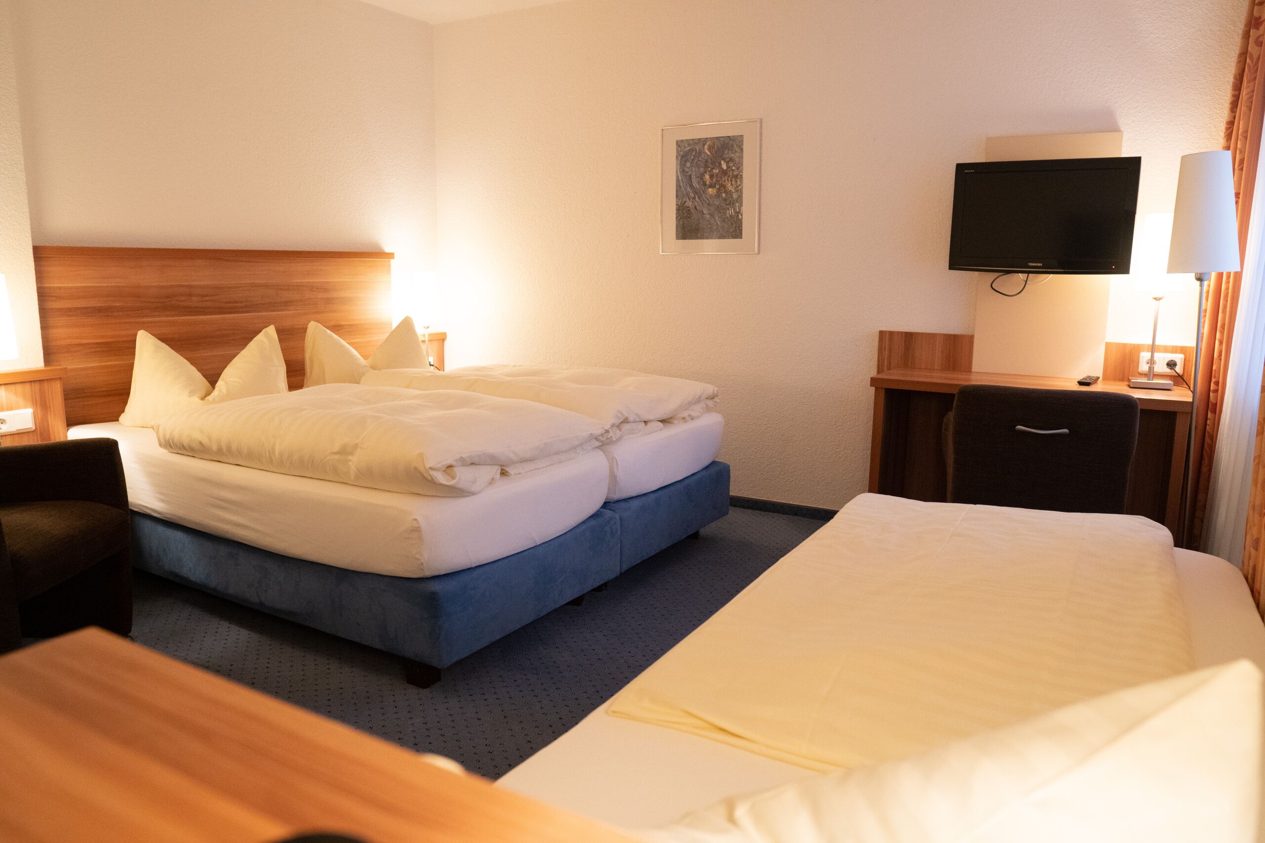 A hotel room with a large bed, a smaller bed, a desk, a chair, a TV, and a lamp.