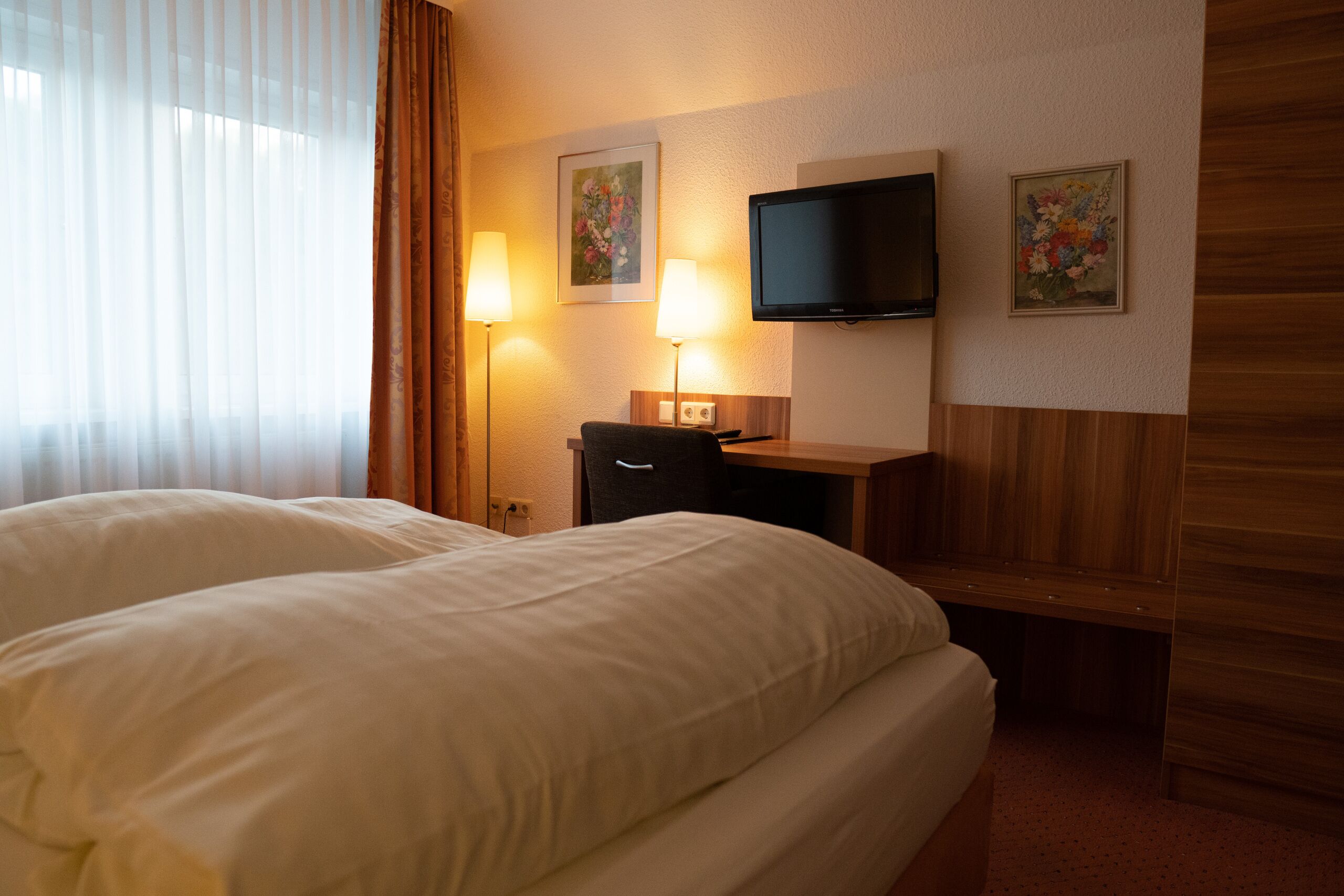 A hotel room with two beds, a desk with a flat-screen TV, two wall-mounted lamps, and floral artwork.