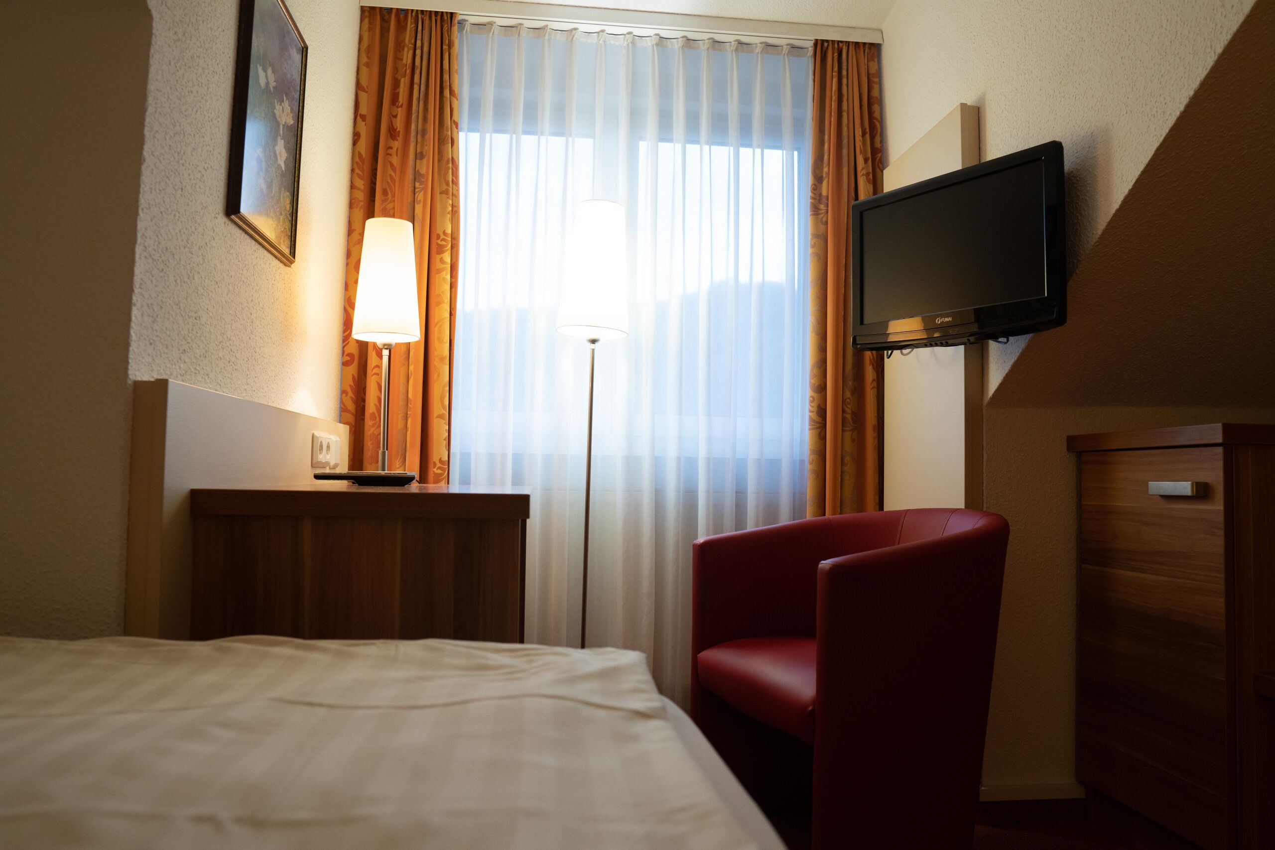 Une chambre d’hôtel avec un lit, un fauteuil rouge, un bureau avec une lampe, un téléviseur fixé au mur et une fenêtre avec des rideaux.