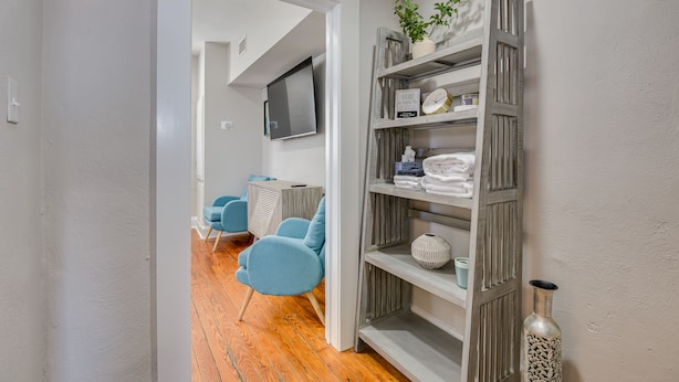Un pasillo con una estantería de madera, un sillón azul y un televisor fijado a la pared.