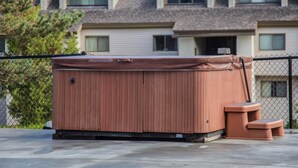 A hot tub with a wooden exterior, a ladder, and steps, located in front of a residential building with a chain-link fence. - Pinnacle F10 (Killington)