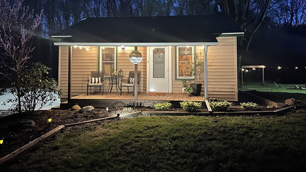 Una pequeña casa con un porche, un letrero que dice "White Oak Cottage", y un área exterior bien iluminada con una mesa y sillas.