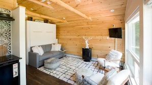 A cozy cabin interior with a wooden ceiling, a gray sofa, a white chair, a fireplace, and a television mounted on the wall.