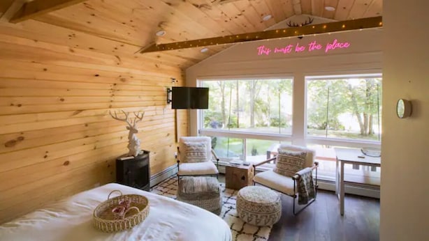 A cozy wooden cabin room with a bed, ottomans, a TV, and a large window offering a view of the outdoors.