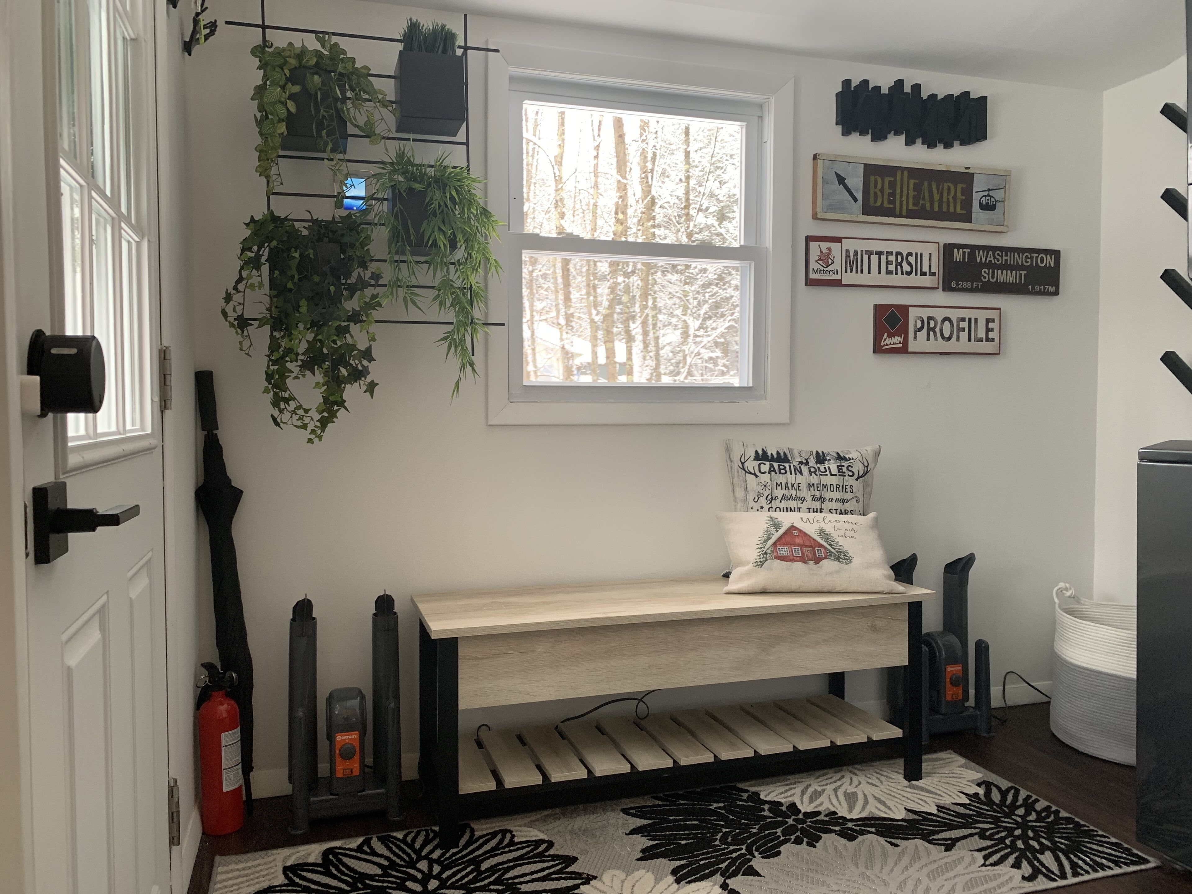 A room with a bed, a window with a view of trees, a mounted plant shelf, and wall decorations including a sign with 'MITTERSILL' and 'MT WASHINGTON SUMMIT' information.