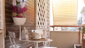 Een klein balkon met een geruit vloertje, witte metalen stoelen, een tafeltje en een decoratieve hangplant.