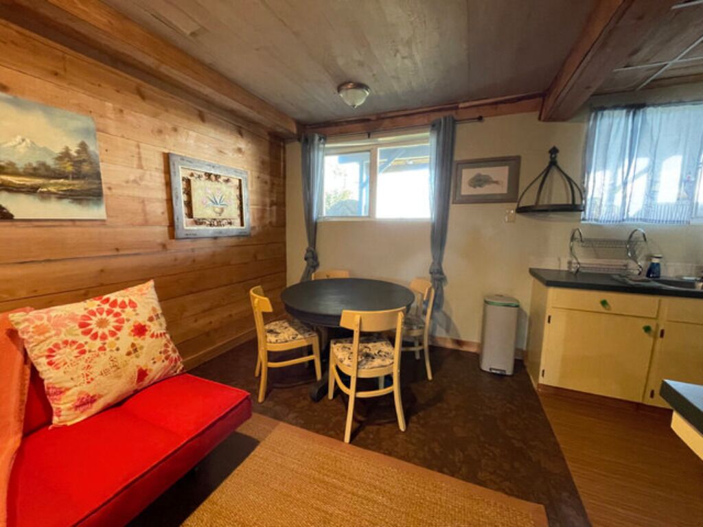 Ett mysigt rum med en röd soffa, ett matbord med stolar, träpanel på väggarna och ett fönster med gardiner.