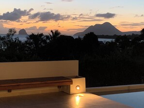 Ein moderner Sitzbereich im Freien mit einer Bank und einem kleinen Tisch, mit Blick auf eine malerische Berg- und Meereslandschaft bei Sonnenuntergang.