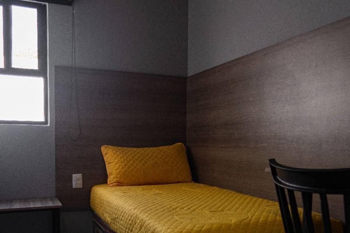 Une petite chambre avec un lit simple, une couverture jaune, un bureau et une chaise. Une fenêtre laisse entrer la lumière naturelle, et un panneau sur le mur indique le point d ’enregistrement.