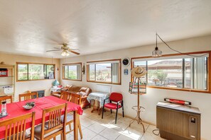 A dining area with a red table, wooden chairs, a ceiling fan, and a view of the outdoors through large windows.