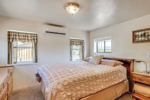 Um quarto com uma cama grande, duas janelas com cortinas, um ar condicionado, uma mesa de cabeceira de madeira com uma luminária e um quadro na parede.