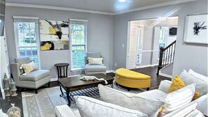 A modern living room with a grey sofa set, a yellow ottoman, and a coffee table. There are two windows with blinds, a staircase in the background, and a painting on the wall. - The Red Magnolia, Refreshing, Cozy, Game Room, Historic Roswell! (Roswell)