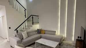 Un soggiorno moderno con un divano componibile, un tavolino e due poltrone. Si intravede sullo sfondo una scala con ringhiere in vetro e un lampadario.