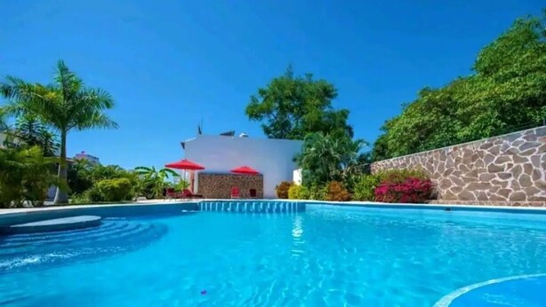 Una piscina con muro in pietra e ombrelloni rossi, circondata dal verde e da un edificio con facciata bianca.