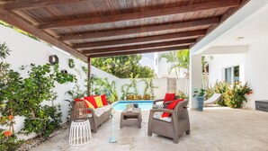A covered patio with wicker furniture, a pool, and lush greenery. - Private villa with pool, enjoy the Dominican Caribbean. (Cabarete)