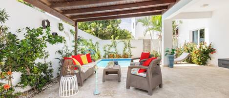 Une terrasse couverte aménagée avec des meubles en osier, une piscine et une végétation luxuriante.