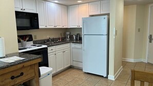 A kitchen with white cabinets, a black microwave, a white refrigerator, a wooden table, and a trash bin.