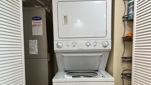A compact laundry room with a front-loading washer and dryer, a Carrier refrigerator, and a utility shelf with cleaning supplies.